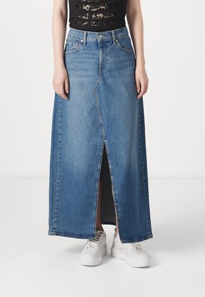 Denim maxi skirt in light blue with a front slit, featuring a classic five-pocket design and a button-fly closure. Worn with white sneakers.