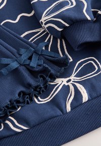 Sweat-shirt bleu marine avec imprimés floraux blancs, ourlet à volants et accents de nœud sur les manches. Confectionné dans un tissu doux et texturé avec des poignets côtelés.