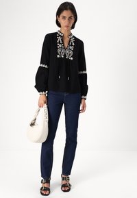Μαύρη μπλούζα με κεντημένο λουλουδάτο σχέδιο στο στήθος και τα μανίκια, συνδυασμένη με μπλε τζιν και μαύρες σαγιονάρες. Το μοντέλο κρατάει μια ελαφριά τσάντα.