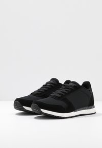Black athletic shoes feature a mix of mesh and suede materials, a textured upper, flat laces, and a white midsole with a speckled black outsole.