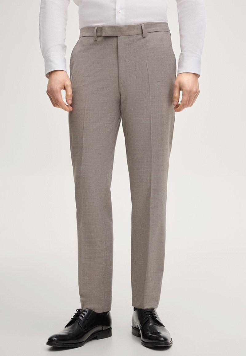 Homme portant un pantalon de costume sur mesure marron clair, des chaussures habillées en cuir noir et une chemise blanche à boutons, debout devant un arrière-plan uni.