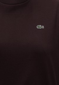 Tissu marron foncé avec un petit logo crocodile vert brodé sur la partie supérieure de la poitrine.