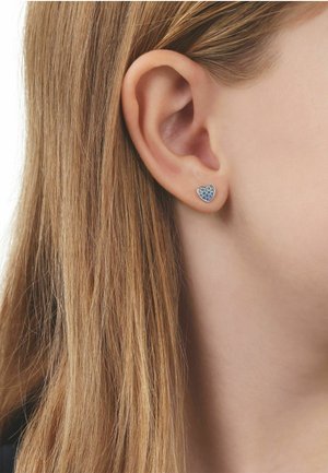 Close-up van het oor van een persoon met lang blond haar, met een kleine zilveren oorbel in de vorm van een hart met blauwe steentjes.