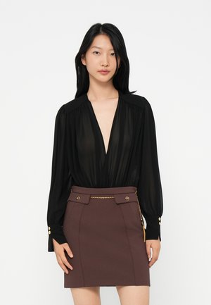 Woman wearing a sheer black long-sleeve blouse with deep V-neck and a brown high-waisted skirt with gold chain detail.