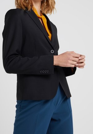 Vrouw draagt een zwarte blazer, mosterdgele blouse en blauwe broek, staand met handen dicht bij elkaar tegen een effen achtergrond.