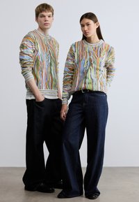 Πλεκτά πουλόβερ με πολύχρωμα αφηρημένα σχέδια, συνδυασμένα με φαρδιά σκούρα τζιν παντελόνια. Η υφή είναι απαλή, αναδεικνύοντας ζωντανές αποχρώσεις και σχέδια.