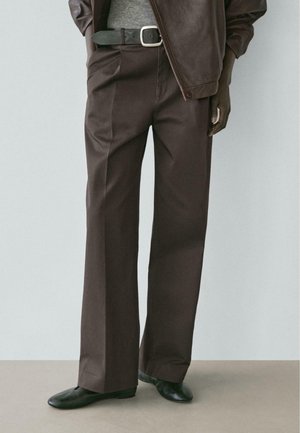 Person iført brune vidde-bukser, sort bælte med sølvspænde, mørkebrun jakke og sorte flade sko, stående på beige gulv.