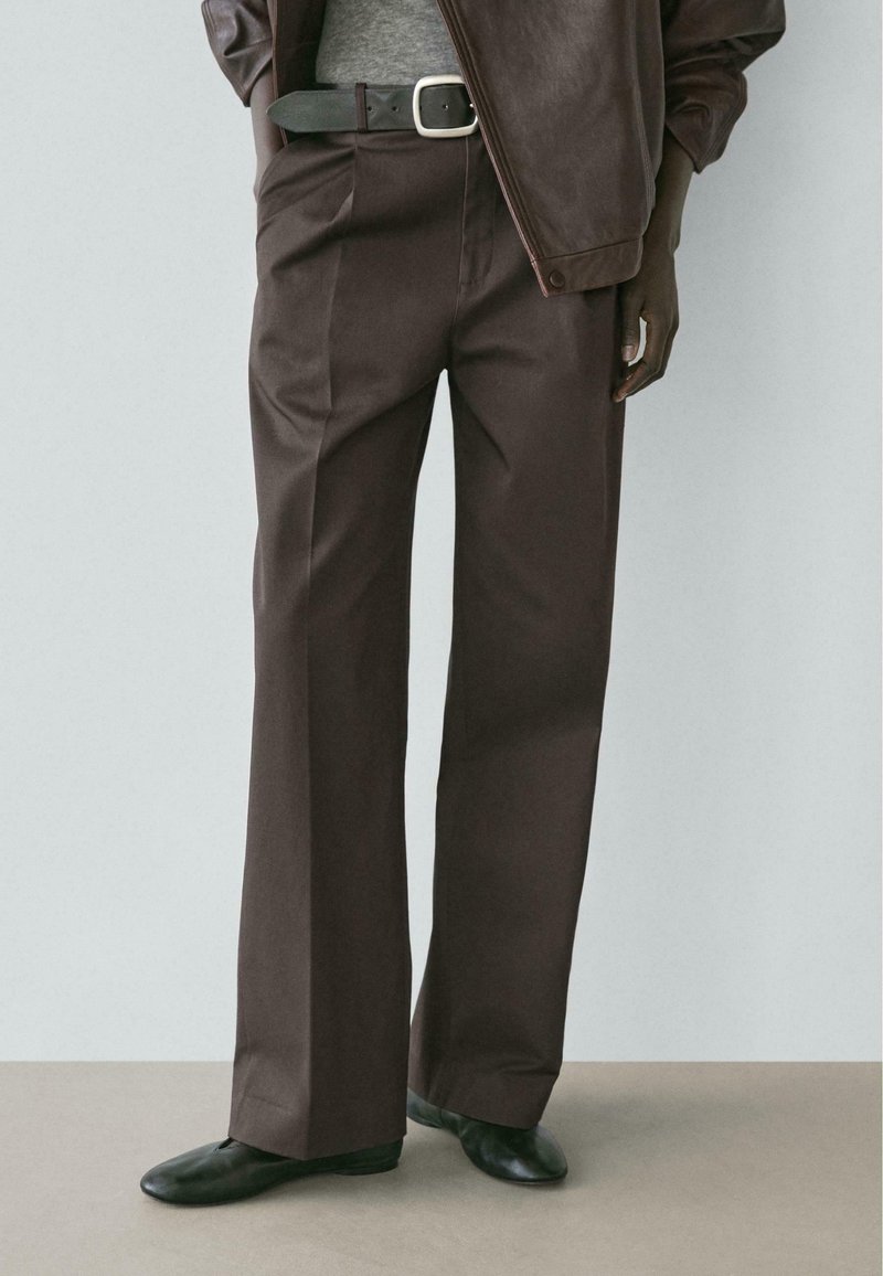 Person trägt braune Hosen mit weitem Bein, schwarzen Gürtel mit silberner Schnalle, dunkelbraune Jacke und schwarze flache Schuhe und steht auf beigem Boden.