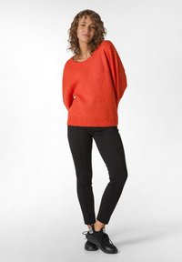 Červený oversized svetr vyrobený z měkké látky, spárovaný s přiléhavými černými kalhotami a černými botami. Svetr má široký výstřih a žebrovaný lem.