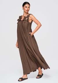 Femme portant une longue robe marron sans manches avec de larges détails floraux sur les bretelles, sandales noires slip-on, posant avec la main dans la poche.