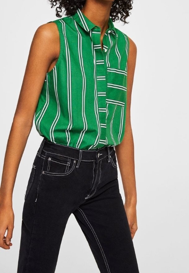 Personne portant un haut vert sans manches avec des rayures blanches verticales rentré dans un jean noir taille haute avec des coutures blanches.