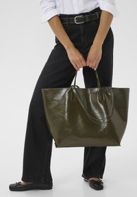 Sac fourre-tout brillant vert olive avec des poignées doubles, de forme rectangulaire et au design minimal, tenu par une personne portant un pantalon et des chaussures noirs.