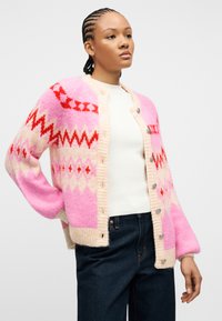 Νέα γυναίκα με πλεξούδες, φορώντας ροζ ζακέτα με σχέδια, λευκή ριμπ πλεκτή μπλούζα και σκούρο μπλε τζιν, στέκεται μπροστά σε απλό φόντο.