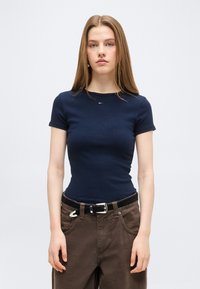 Νέα γυναίκα με μακριά μαλλιά, φορώντας σφιχτό μπλε μπλουζάκι με κοντά μανίκια και φαρδιά καφέ παντελόνια με μαύρη ζώνη, στέκεται μπροστά σε ένα απλό φόντο.