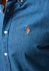 Knopenblouse van denim in medium blauw met een gladde textuur. Beschikt over witte knopen en een geborduurd logo van een ruiter in levendige kleuren.