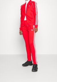 Survêtement de sport rouge avec des rayures blanches sur les côtés, composé d'une veste à fermeture éclair et d'un pantalon à taille élastique, associé à des baskets noires.