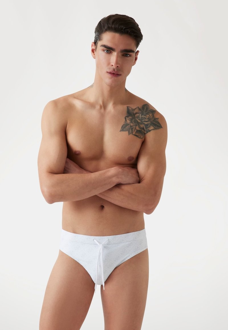 Witte zwemslip met een subtiel patroon, koord in de taille en een soepele textuur; model met gekruiste armen, die een gespierd figuur showt.