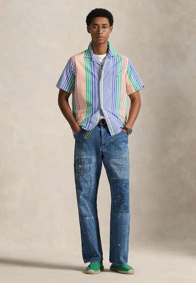 Πολύχρωμο πουκάμισο με κοντά μανίκια και κάθετες ρίγες, σε συνδυασμό με σκισμένο τζιν μπλε χρώματος που διαθέτει τσέπες cargo και πιτσιλιές μπογιάς. Πράσινα παπούτσια.