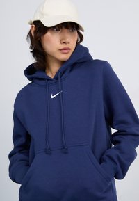 Mørkeblå hettegenser i bomullsblanding, med kengurulomme og hvit Nike-logo. Akkompagnert av en lyse farget caps.