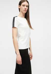 Γυναίκα με μακριά καστανά μαλλιά που φορεί λευκή κοντομάνικη μπλούζα με μαύρες λωρίδες στα πλάγια και μαύρη φούστα, στέκεται μπροστά σε απλό λευκό φόντο.