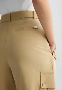 Pantalons cargo beiges avec une texture lisse, présentant une poche arrière avec un accent argenté et une poche latérale fermée par un bouton.