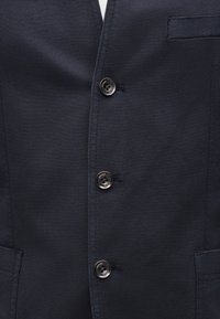 Σκούρο μπλε σακάκι με τρία μαύρα κουμπιά με την επιγραφή "JOOP! JEANS" και μια μικρή τσέπη στο στήθος στη δεξιά πλευρά.