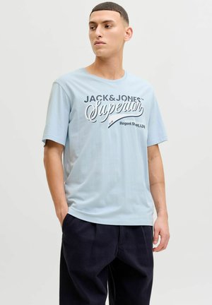 Light blue cotton t-shirt featuring a printed logo in dark navy, short sleeves, and a relaxed fit, paired with dark trousers.