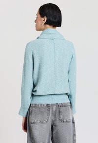 Hellblauer Pullover mit hohem Kragen, strukturiertem Strickmuster und geripptem Saum. Getragen mit grauen, hoch taillierten, lässigen Jeans.