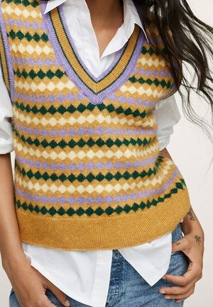Femme portant un gilet tricoté à motif losanges jaune, vert et lavande sur une chemise blanche à manches retroussées et un jean bleu.
