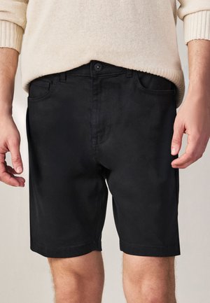 Hombre con pantalones cortos negros con botones y un suéter beige, de pie con los brazos relajados a los lados contra un fondo liso.