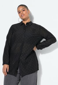 Schwarze, knöpfbare Hemd mit langen Ärmeln, ausgestattet mit einem strukturierten Stoff und kleinen weißen Punkte-Mustern. Trägt es über grauen Hosen.