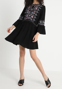 Vestido negro con bordados florales en rosa y azul. Presenta mangas largas y acampanadas y un dobladillo con volantes. Se lleva con mules negros de tacón.