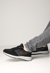 Scarpe sportive nere e grigie con tomaia in suede e tessuto, dotate di una suola ammortizzata e un logo sul lato. Indossate con jeans grigi.