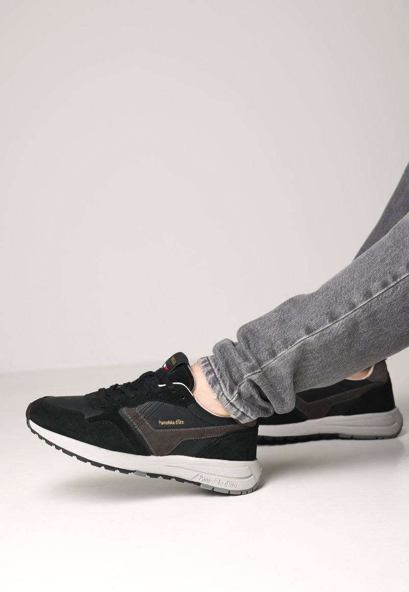 Scarpe sportive nere e grigie con tomaia in suede e tessuto, dotate di una suola ammortizzata e un logo sul lato. Indossate con jeans grigi.