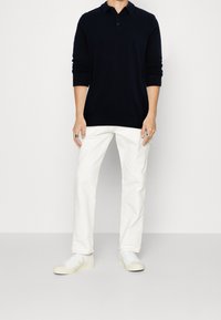 Man i en svart långärmad pikétröja, vita byxor, vita sneakers och silverringar mot en enkel vit bakgrund.