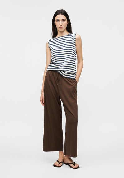Femme portant un haut sans manches rayé bleu marine et blanc, un pantalon marron ample avec cordon de serrage, et des tongs noires, debout devant un fond blanc.