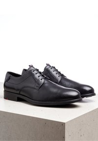 Chaussures habillées en cuir noir avec un design épuré, bouts ronds et lacets. Elles présentent un petit accent au niveau du talon et une semelle à motif standard.