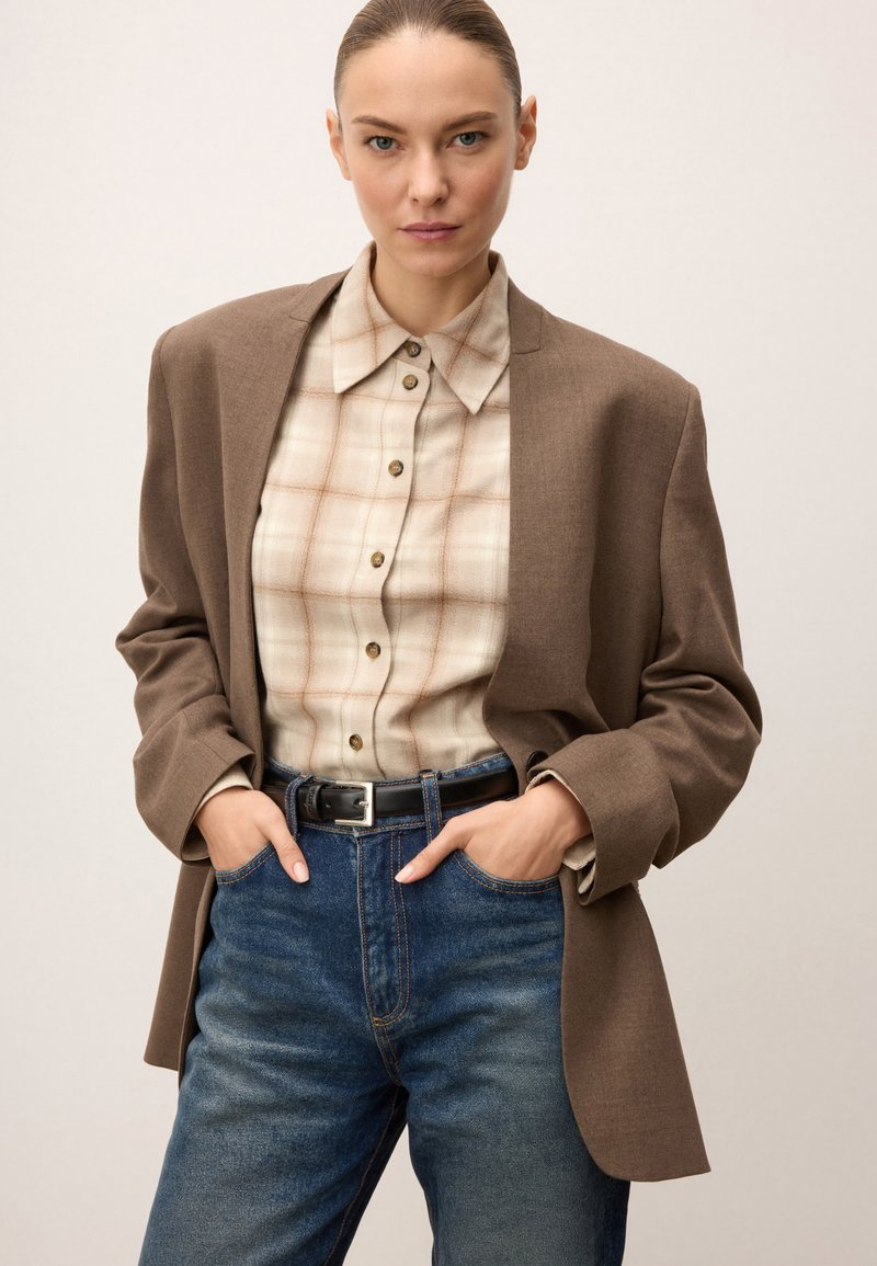 Barna blazer bézs kockás inggel, gombos részletekkel, kék farmernadrággal párosítva. Az öltözethez fekete öv tartozik ezüst csattal.