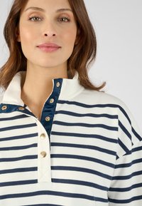 Camisa polo a rayas en blanco y marino, con un cuello alto y botones dorados. Tejido suave con un corte relajado, que presenta una tapeta de botones.