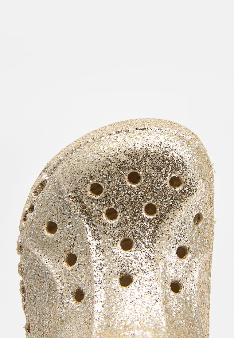 Zoccoli dorati glitterati con fori di ventilazione, punta arrotondata e texture liscia e lucida. Materiale leggero con un design imbottito.