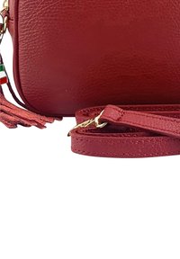 Rote Ledertasche mit abnehmbarem Schulterriemen und Quaste, ausgestattet mit einem kleinen Emblem in den Farben Grün, Weiß und Rot.