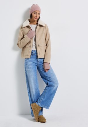 Donna che indossa una giacca di shearling beige, un cappello di maglia rosa e guanti, jeans a gamba larga di un blu chiaro e stivali alla caviglia in suede color sabbia, in piedi contro un muro bianco.
