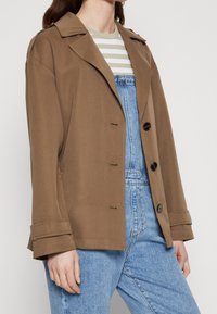 Hnědá, oversized bunda s klopovým límcem, jednoramenným zapínáním a velkými černými knoflíky, v kombinaci s pruhovaným topem a džínovými overaly.