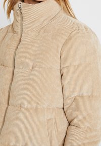 Beige corduroy pufferjakke med høj krave, der har ribbet tekstur, lynlås lukning og polstrede sektioner til isolering.