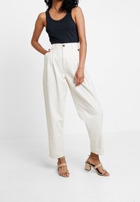 White high-waisted pleated trousers with two buttons, paired with a black tank top and beige block-heeled sandals.
