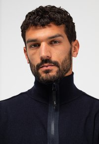 Marineblauer Pulli mit hohem Kragen, durchgehendem Reißverschluss und strukturiertem Stoff. Hat ein feines Strickmuster und dezente Markenkennzeichnung am Reißverschluss.