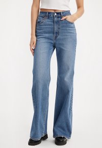 Femme portant un jean bleu taille haute à jambes larges et des chaussures noires, avec une main dans la poche et un top court blanc.