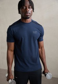 Navy short-sleeve athletic shirt with black shoulder accents and EA7 logo, paired with black pants; holding chrome dumbbells. Smooth fabric.