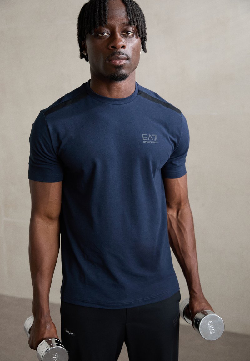 Navy short-sleeve athletic shirt with black shoulder accents and EA7 logo, paired with black pants; holding chrome dumbbells. Smooth fabric.