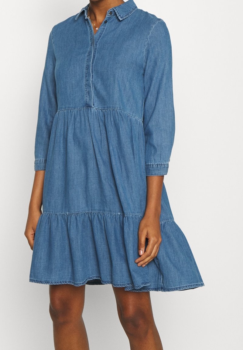 Robe en jean bleue longueur genou avec manches longues, col et jupe à volants portée par une personne à la peau foncée.